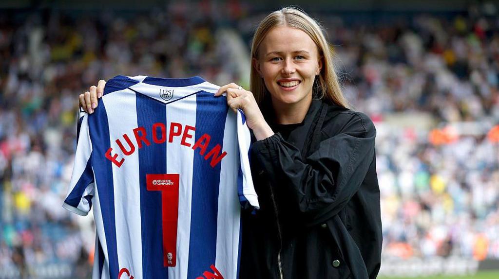 Hannah Hampton holds up a blue and white striped West Bromwich Albion shirt on the pitch at The Hawthorns, with a crowd blurred in the background. The shirt has a number 1 on the back and European written. She is wearing a black jacket and black t-shirt and smiling.