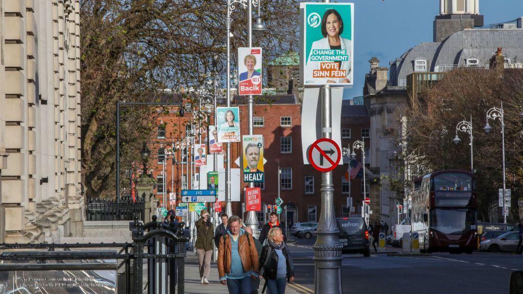 Pedestrians walk along a street in Dublin which is covered in election posters along the lampposts