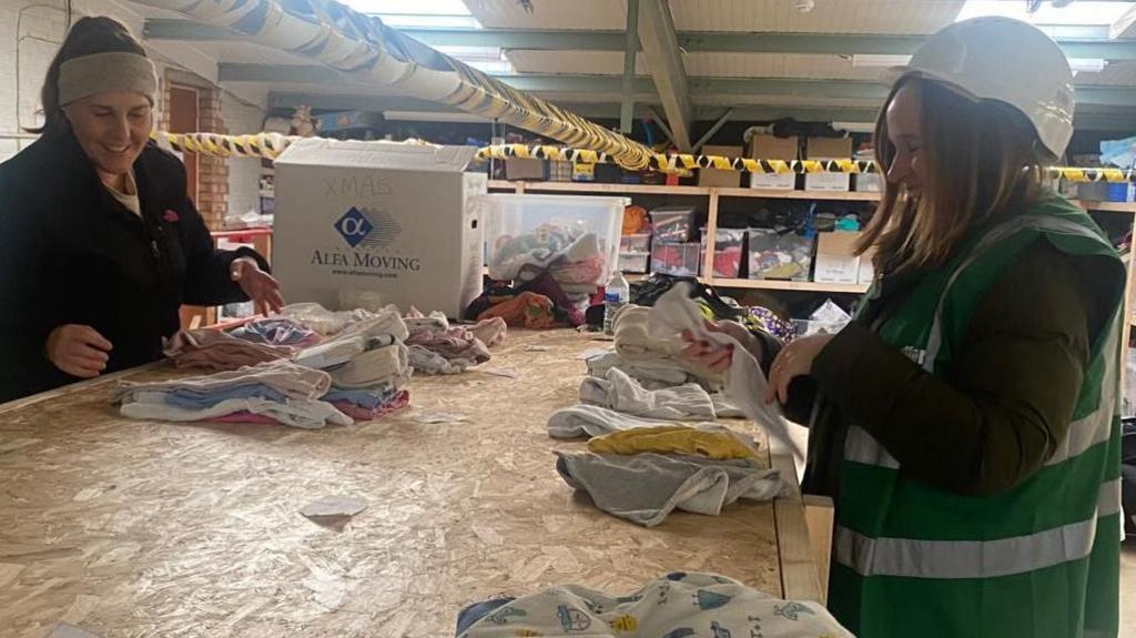 Two women inside a large industrial unit, sorting baby clothes out. The woman on the right is wearing a hard-hat with a green high vis jacket and the woman on the left is hearing a head band, and a dark top. They are both smiling. There are baby items on a wooden desk and rows of boxes on shelves behind. 