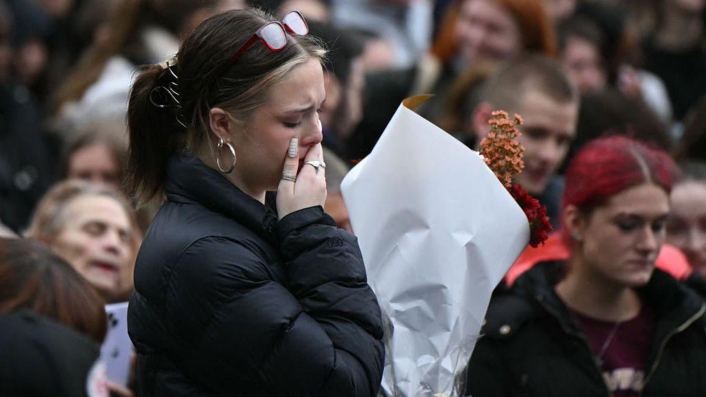 A woman in a black puffer jack holds her hand to her mouth, visibly upset and holding orange and red flowers. A crowd is stood in the background.