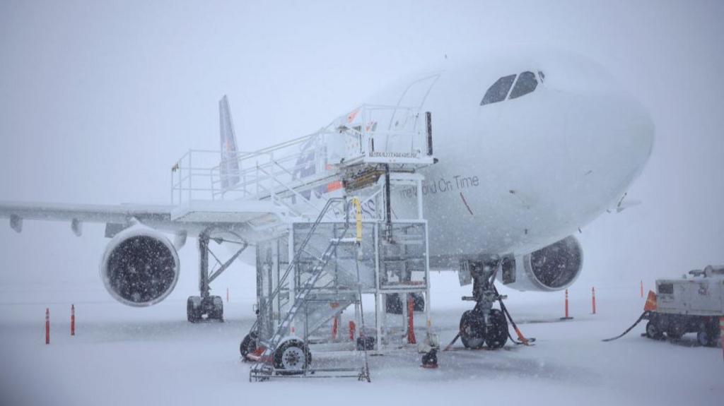 FedEx plane sits on runway covered in snow with snow falling and scaffolding resting up against the plane