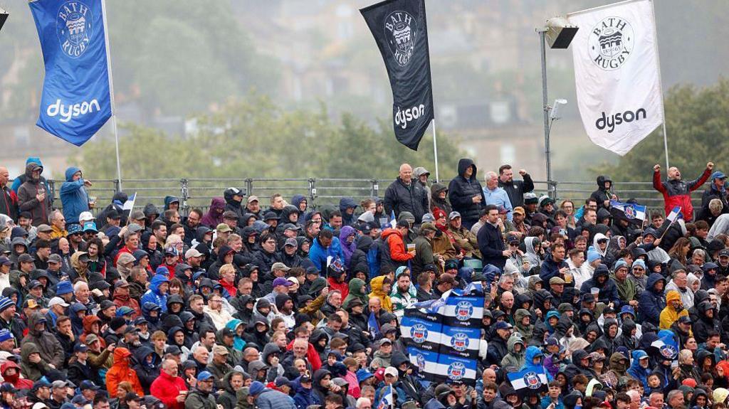 A packed grandstand at The Rec, the home of Bath Rugby club. Fans can be seen wearing blue lack and white and waving flags of the club. Building from the city of Bath can be seen in the background.