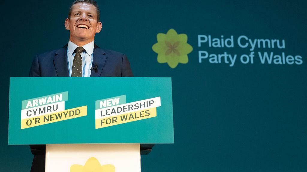 Rhun ap Iorwerth smiling during his Plaid Cymru conference speech in Swansea, standing at the podium.