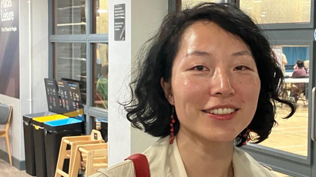 Yuan Yang with short, black, wavy hair smiling at the camera with an election count visible through the window behind her