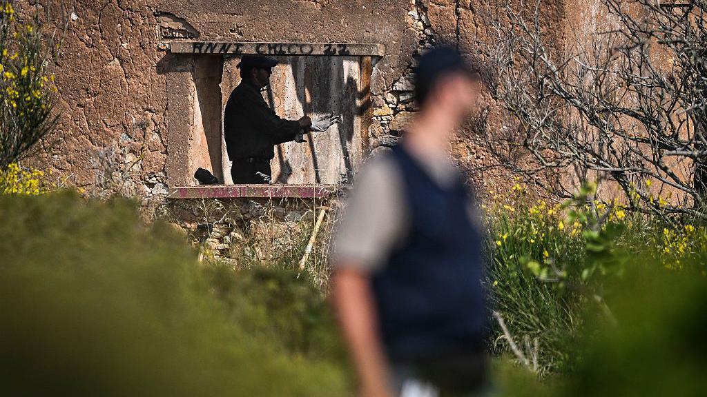 Derelict building with a man standing inside it, visible through the hole in the wall where a window would have been. On the upper frame is black graffiti. A blurred man is standing in the foreground to the centre-right of the picture. There is vegetation surrounding the building.