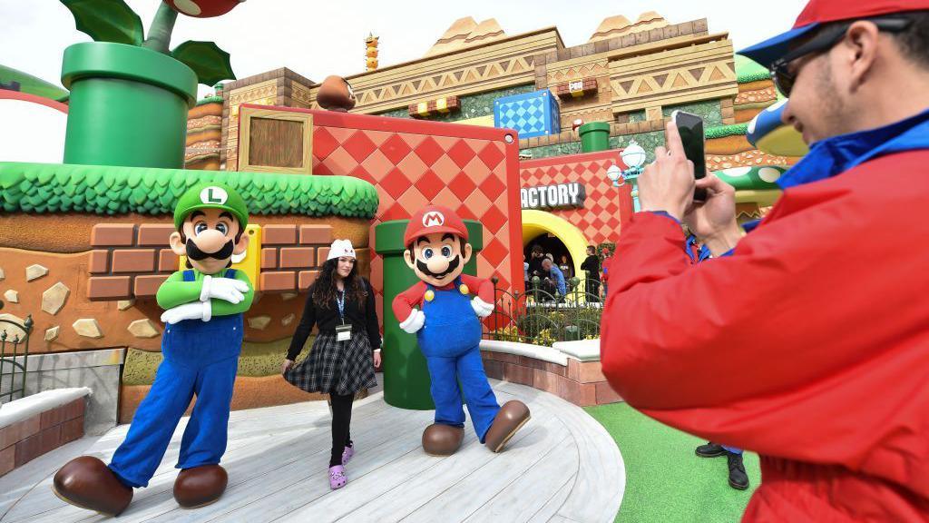 A member of staff takes a photo of a member of the public posing with actors dressed as Mario and Luigi at a Universal theme park