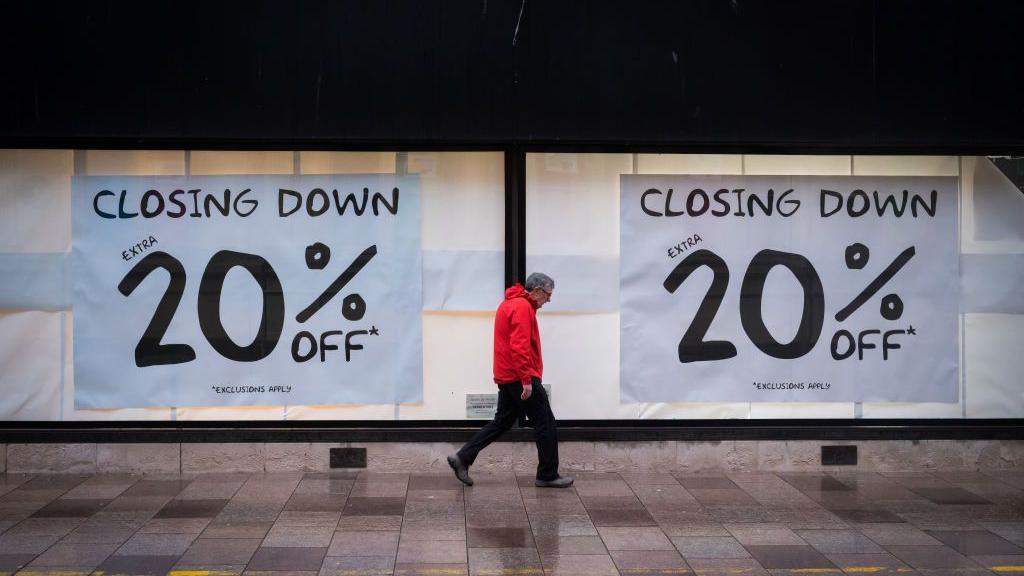 A man in a red coat walks past a shopfront with windows displaying large posters, the text on the posters reads: "CLOSING DOWN, EXTRA 20% OFF"