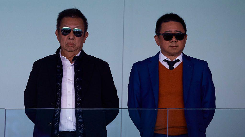 Two men, wearing sunglasses, look ahead behind a glass barrier at a football stadium.