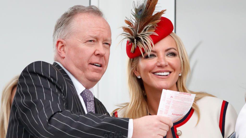 Doug Barrowman, a man with greying hair, is wearing a grey chalk-stripe suit and white tie with purple geometric pattered shirt. He is holding a number of betting slips. Michelle Mone is in a cream, red and black jacket with matching hat. She has blonde hair and is smiling.