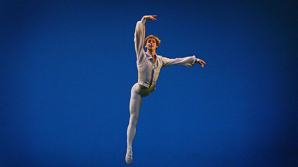 Russian ballet star Vladimir Shklyarov poses in a ballet pose, wearing a white and gold costume. His head is tilted back and his eyes are closed. The background is dark blue.