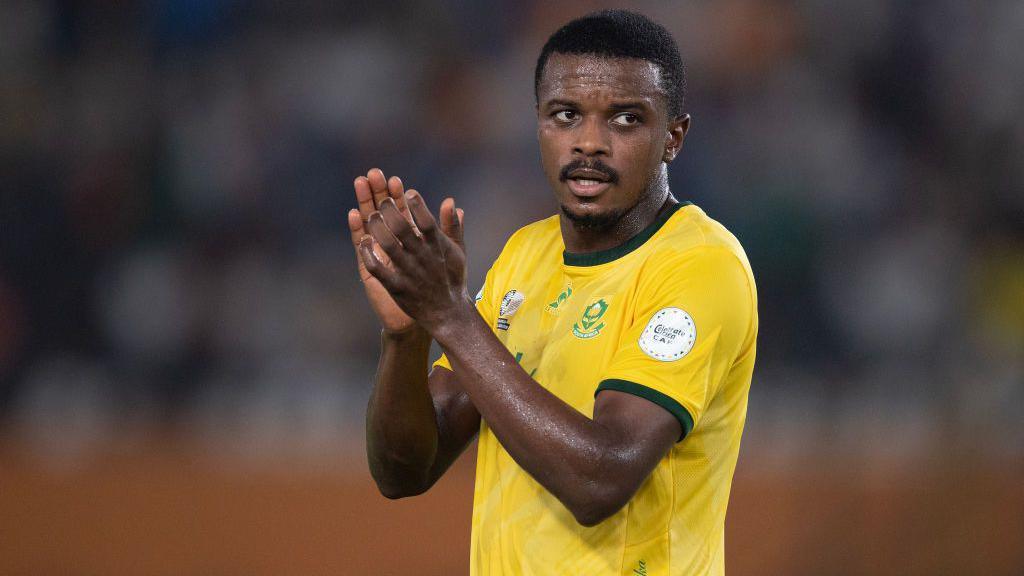 Teboho Mokoena, pictured from waist up in the traditional South African gold jersey, claps his hands as he is subbed off during a football match