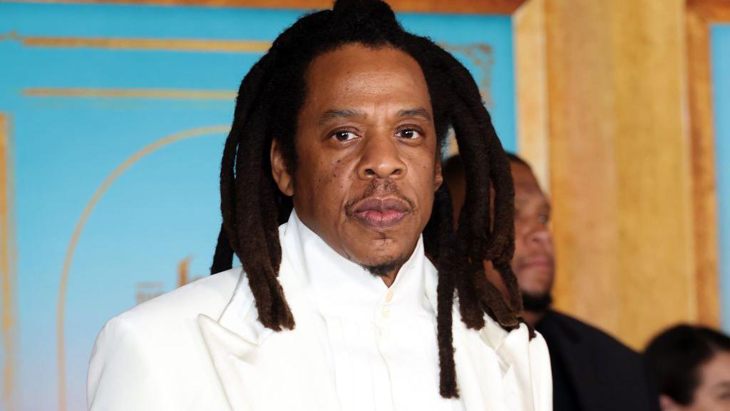 Jay-Z in a white suit at a party. He has a neutral facial expression. 