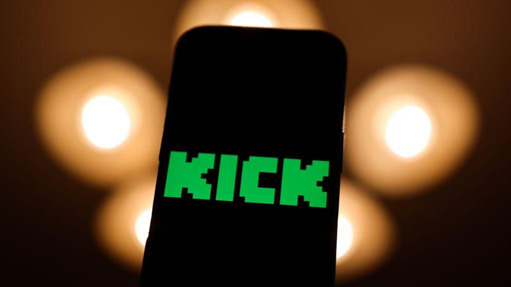 A phone screen with the Kick logo in green on it. Black background with orange blurry lights. 