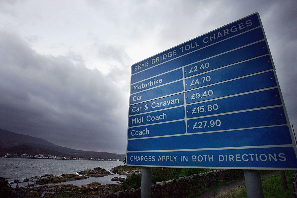 A blue and white road sign listing the charges for crossing the Skye Bridge for different vehicles. It says it costs motorbikes £2.40, cars are £4.70, car and caravans are £9.40, "midi coach" is £15.80 and a coach is £27.90. It says charges apply in both directions. Behind the sign is a large expanse of water and an island with white housing behind it.