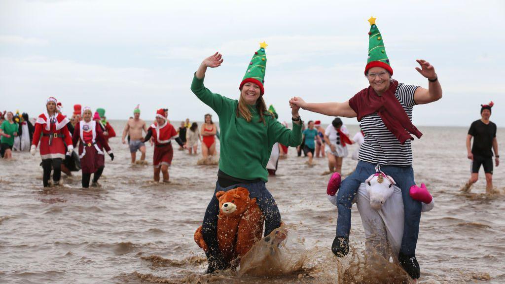 A group of people in the sea wearing Christmas hats and costumes. At the forefront of the image are two women wearing Christmas tree hats and a bear and unicorn suit. 