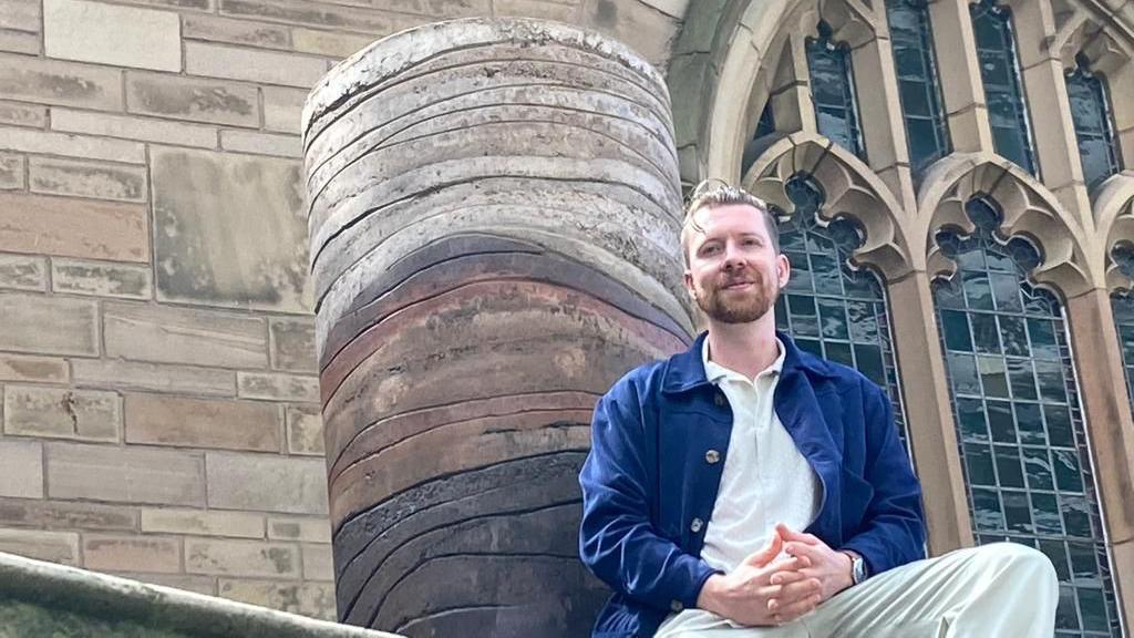 Benjamin Stephenson is wearing beige trousers and a blue jacket over a white shirt. He is sat at the base of his sculpture which is a cylinder showing layers of brown compacted soil and clay.