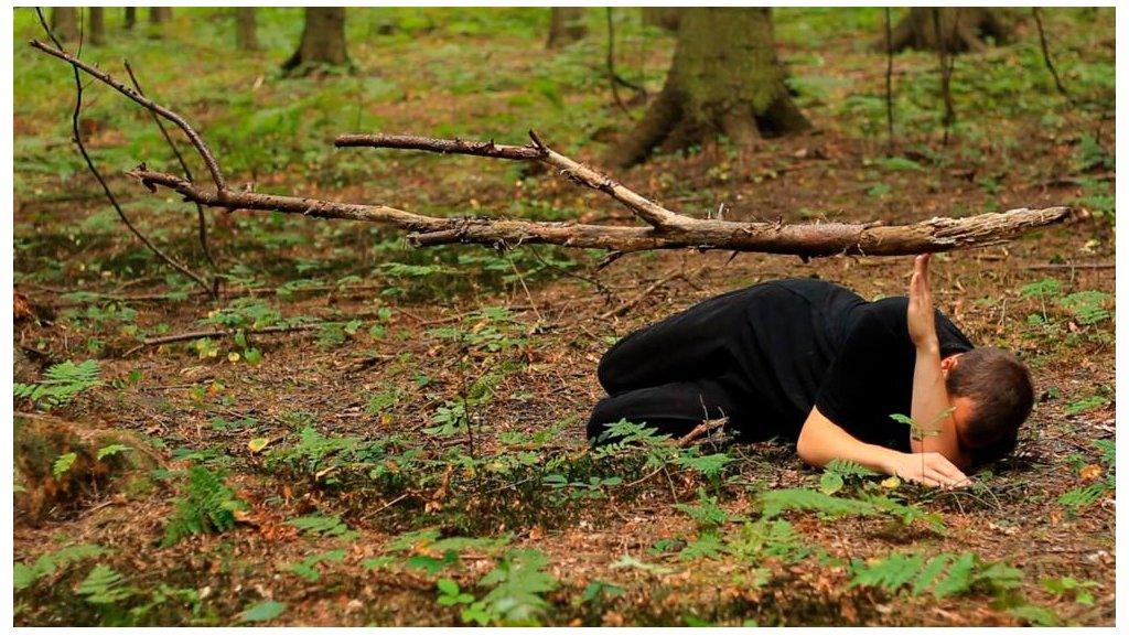 Man dressed in black curled up on the ground and holding a branch with the tips of his finger.