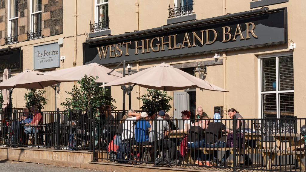 Front entrance of West Highland Bar in Portree, Isle of Skye. The venue has a black and gold sign with a number of tables, umbrellas and patrons outside on a sunny day. 