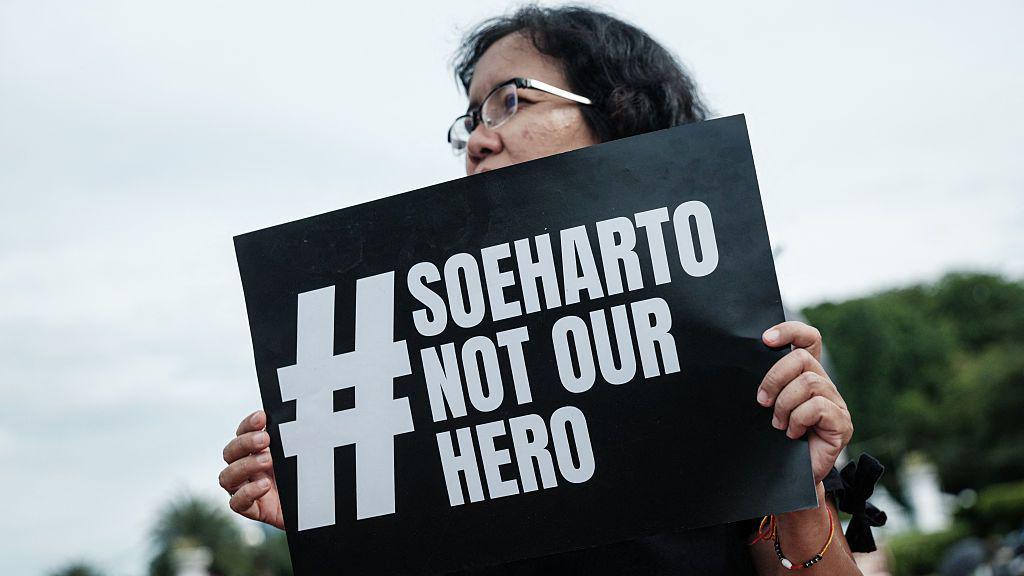A woman holding up a black sign that reads: Soeharto not our hero.