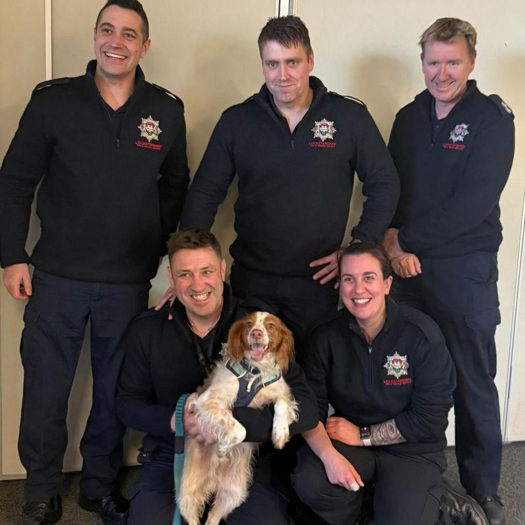 Five firefighters grinning in uniform as they cuddle up with a dog. Three of them, men, are standing, while a man and a woman kneels. The kneeling man holds a dog with brown ears and brown/white fur on the rest of its body with a green harness around its neck and torso.