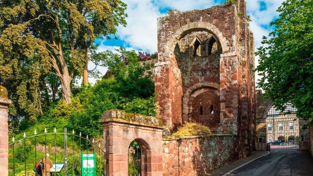 Entrance to Rougemount Castle, Exeter, Devon, England. It is a large ruin of a red stone structure, with a newer black gate to the left hand side.