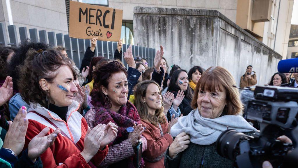 Gisele Pelictor, a woman with auburn hair in a bob, wearing a coat and a pale blue scarf, surrounded by a crown of women on one side applauding her, some who seem overcome with emotion, and one person holding a sign saying, "Merci Gisele". On her other side is a TV camera.