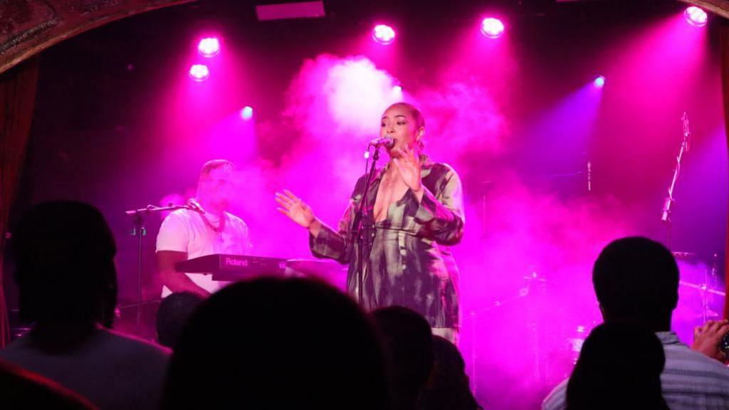 A woman bathed in purple light sings into a microphone while holding up both her hands on stage. A keyboard player can be seen to her left and a crowd of people watching in the foreground. 