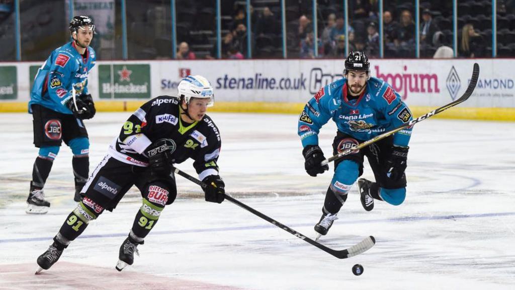 Two blue uniformed ice hockey players skate towards a black uniformed player. A small crowd can be seen in the background behind a glass divider.  