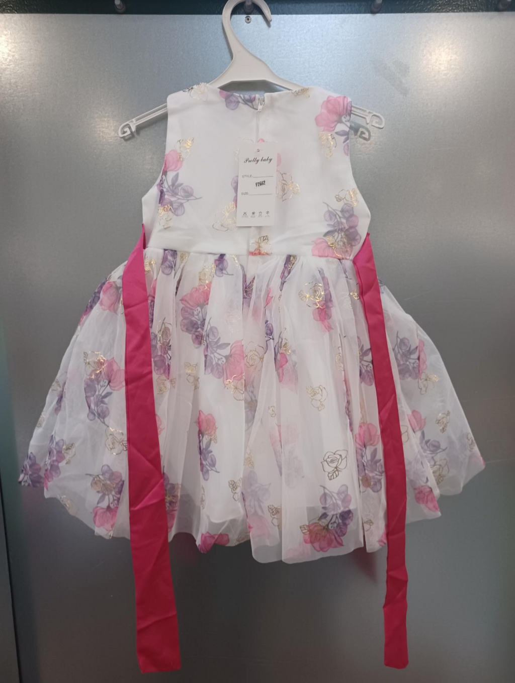 White children's dress with a pink and purple floral pattern and chiffon skirt. The dress is hanging on white plastic clothes hanger against a silver metal backdrop. The dress has a dark pink tie-at-the-waist ribbon which is untied and appears to be longer in length than the dress itself.