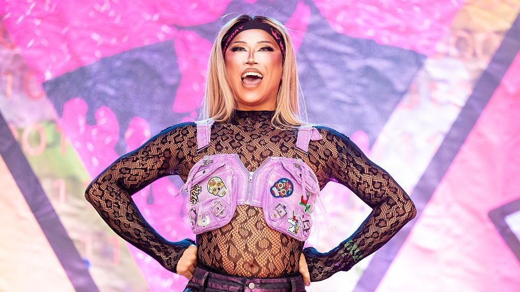 A drag performer in a fishnet, leopard-print top stands on stage, hands on hips, smiling. They're wearing a pink, PVC, life jacket-style top with sugar skull patches on it.