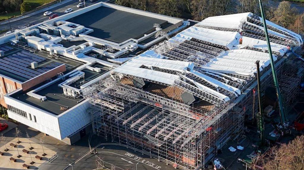 Aerial view of the Sands Centre taken in January after the storm. The right-hand side of the venue is covered with scaffolding, as is the section of roof above. Parts of a temporary canopy appear to have blown off. To the front and side of the building is a car park, while fields, a river and trees can be seen in the background.