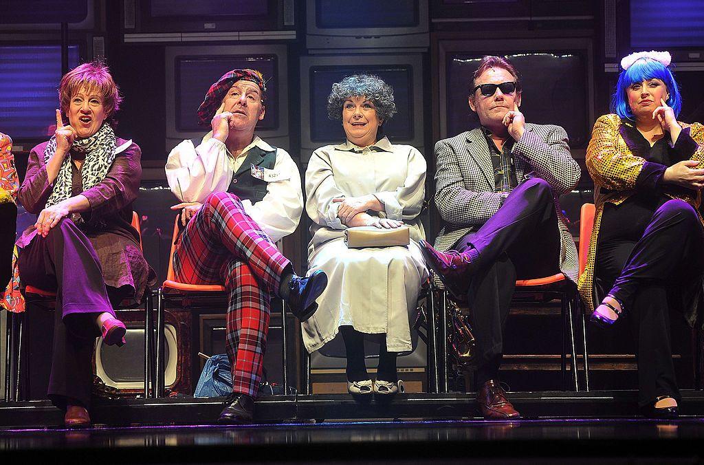 Elaine C Smith in a beige coat, with grey, curly hair, seated on stage between four people dressed in more outlandish outfits