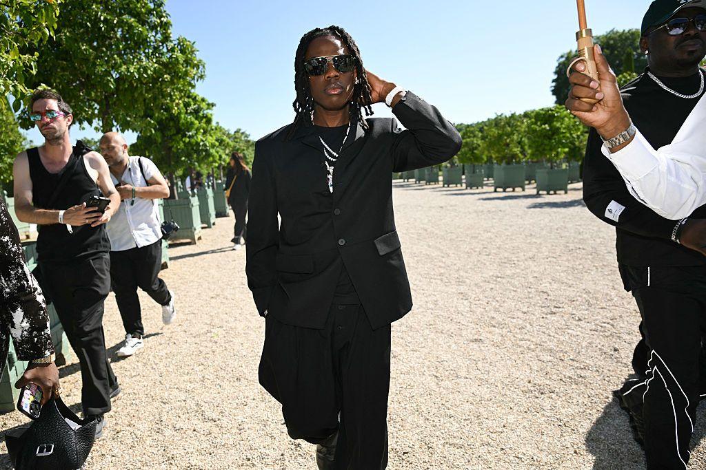A man dressed in black walks through the Orangery of the Palace of Versailles. He is flanked by an entourage.