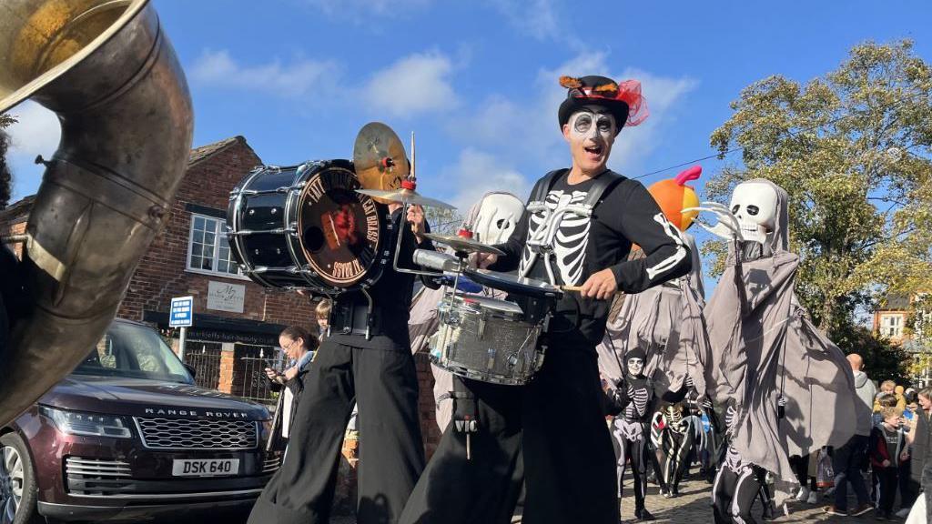 Multiple skeleton characters playing instruments, wandering through the streets of Lincoln on stilts. 