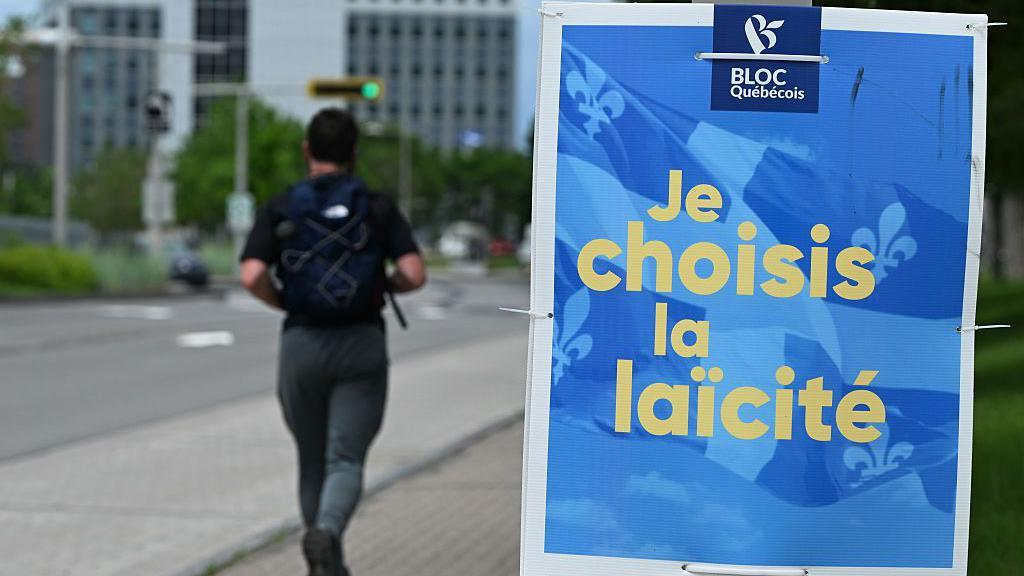 A man is black athletic clothes runs by a blue sign for the federal Bloc Québécois that reads in French 'I choose secularism'