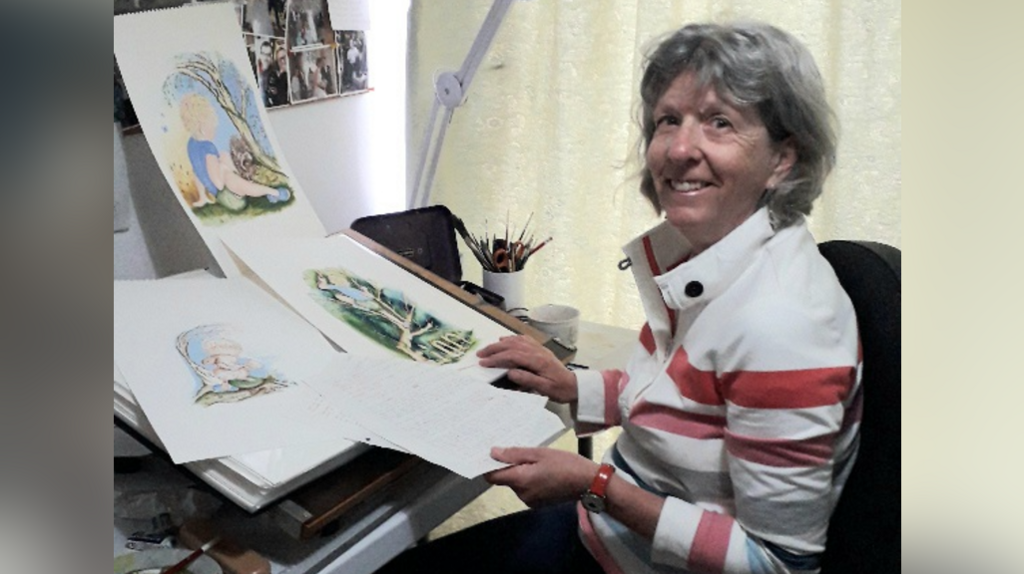 Jan sat at a desk. She has large illustrations for a children's book in front of her. She is looking over her shoulder and smiling. She is wearing a white, pink and grey striped jacket and has short grey hair.