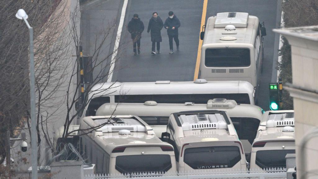 Three people in black walking along a road in the presidential compound, with several buses parked right in front of the gate