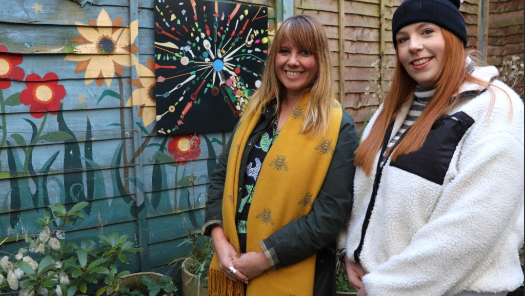 Prof Claire Gwinnett and Laura Wilkinson are stood outside by a green painted fence next to artwork. Claire has long brown/blonde hair and is wearing a large yellow scarf over a dark jacket and patterned top. Laura has a white jacket on over a top with long, red hair and is wearing a bobble hat. 
