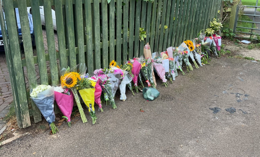 Floral tributes in a line against a fence
 