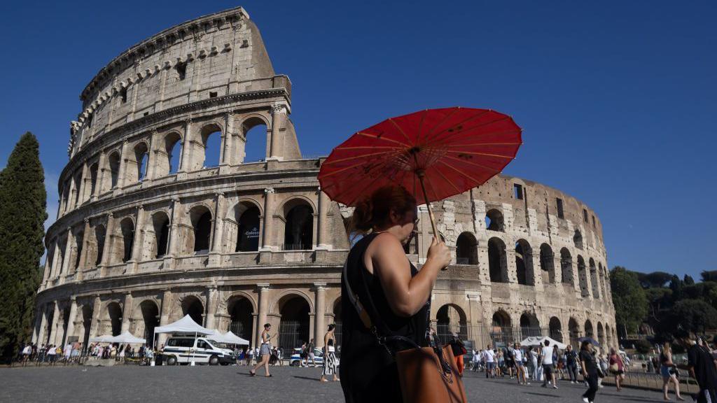 A woman walks past a historical building while shielding herself from the sun with an umbrella