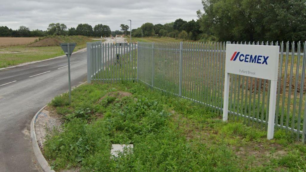 The entrance to a concrete plant. A road passes by a silver metal fence next to a white sign that says "Cemex - Pyford Brook".