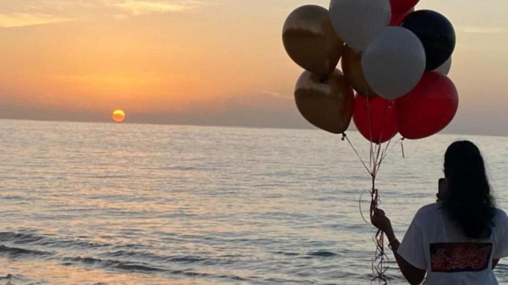 A woman wearing a t-shirt has her back to the camera. She is holding a cluster of balloons in her left hand. In front of her is the sea and the sun can be seen setting along the skyline