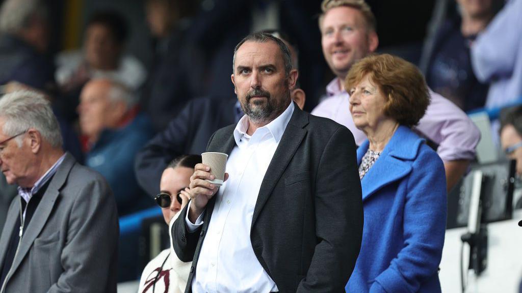 A man in a white shirt and black blazer holds a hot drink whilst watching a football match in a crowd of people. 