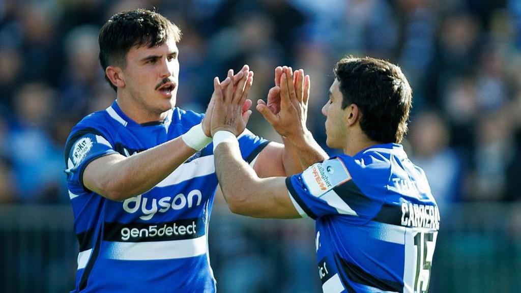 Ewan Richards (left) holds both hands up with palms wide to clap with Santiago Carreras (right)