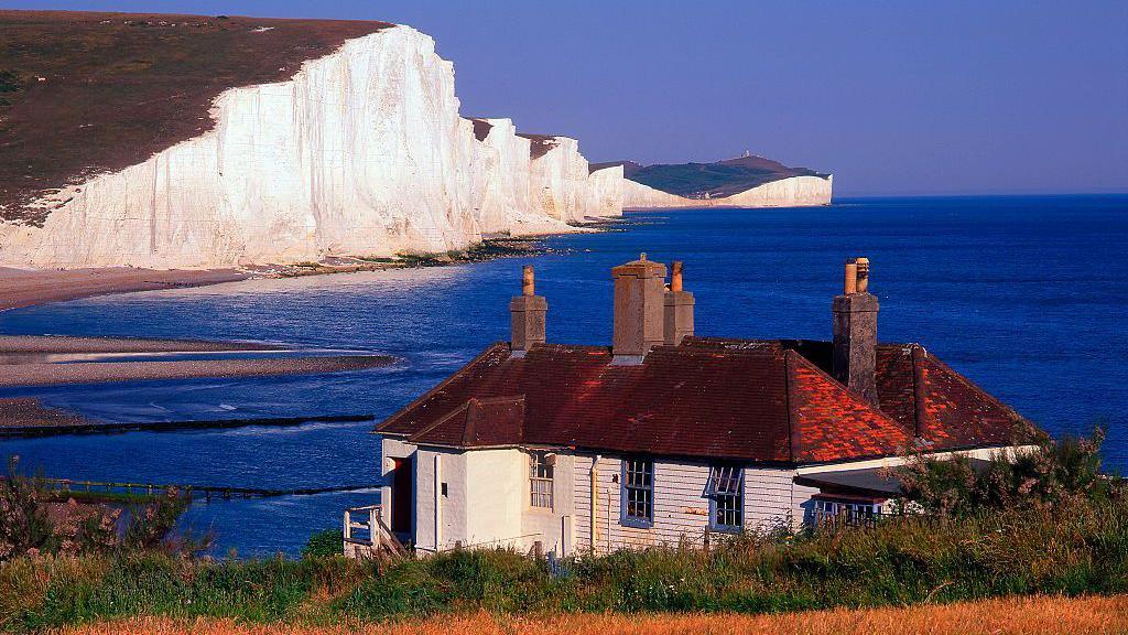 The Seven Sisters cliffs stretching in to the distance with a red-roofed cottage in the foreground and the blue sky and sea visible into the distance.