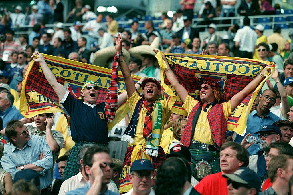 Three fans holding Scotland flags and singing their hearts out in the Stade de France - they wear kilts and Scotland tops and also wear Scotland and Brazil scarves.