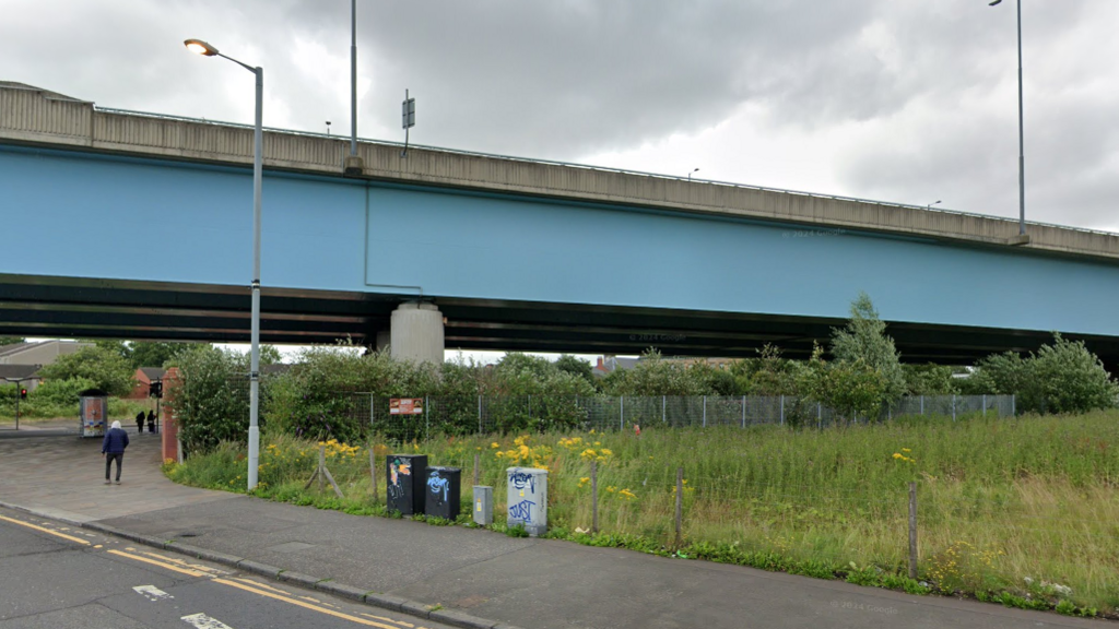 View or grassy area with lampposts and fence under large metal and concrete motorway flyover.