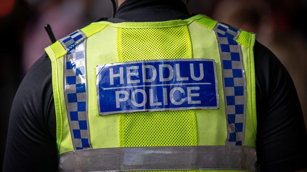 The back of a police officer in Wales. He is wearing a neon yellow vest over a black top. The vest has a blue box with grey writing with 'Heddlu Police'