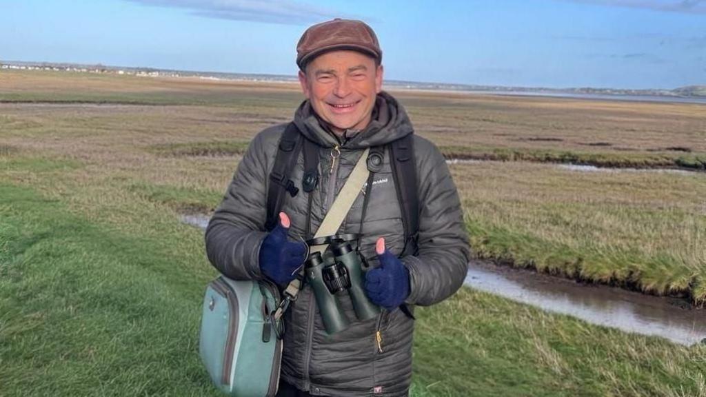 Simon smiles as he holds binoculars in his hands. He stands in front of a green nature reserve. Simon wears a grey jacket, a blue shoulder bag, navy gloves and a brown cap.
