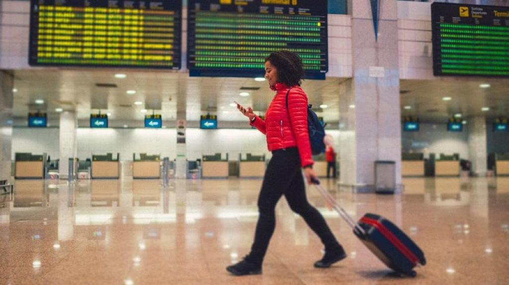 Young woman with wheeled luggage walking at the airport and texting